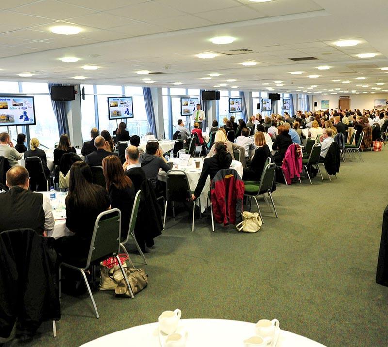 2 rows of tables with business guests listening to a standing speaker.